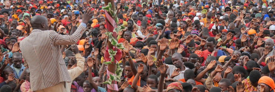 View from a stage, we see the back of a preacher raises his hands on a stage in front of hundreds of Burundians also raising their hands with eyes closed